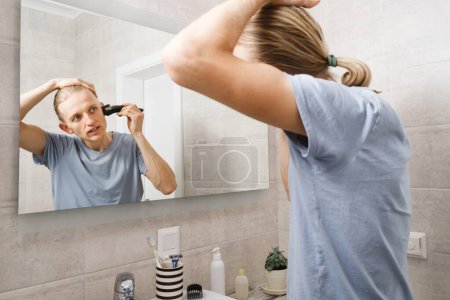 Téléchargez les photos : Hygiène personnelle, homme caucasien couper ses propres cheveux dans la salle de bain avec rasoir électrique sans fil devant le miroir - en image libre de droit