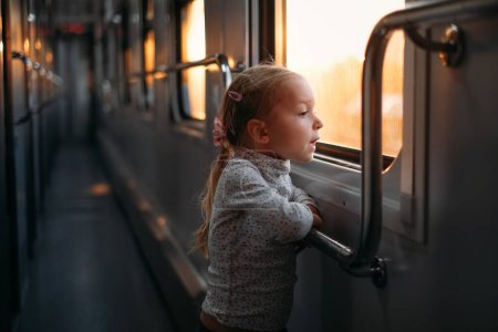 Kleinkind beim Blick durch das Zugfenster bei Sonnenuntergang, helles Sonnenlicht, stimmungsvolle Bahnfahrt mit Kindern. Mädchen glücklich, den Weg am Abend zu erkunden. Spannender Familienausflug