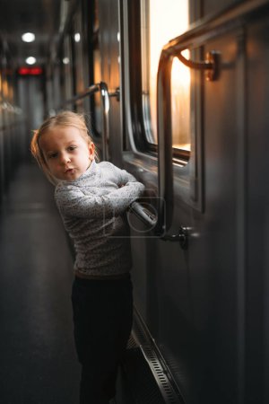 Toddler child looking though train window on sunset, bright sunlight, atmospheric travel by railway with kids. Girl happy exploring the way in evening. Exciting family trip