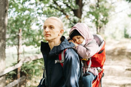 Photo for Man hiking with sleepy toddler child in the mountain. Kid sitting in backpack on father back. Active family adventure in forest with tired girl. Cold weather trip, Nordic walk tour - Royalty Free Image
