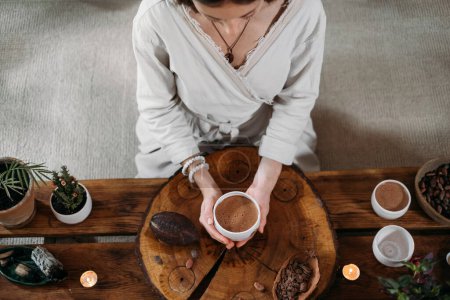 Photo for Hot handmade ceremonial cacao in white cup. Woman hands holding craft cocoa, top view on wooden table. Organic healthy chocolate drink prepared from beans, no sugar. Giving cup on ceremony, cozy cafe - Royalty Free Image