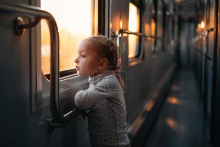 Toddler child looking though train window on sunset, bright sunlight, atmospheric travel by railway with kids. Girl happy exploring the way in evening. Exciting family trip