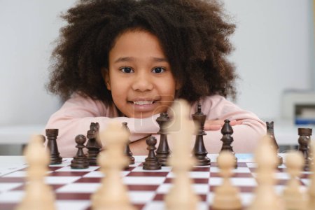 Téléchargez les photos : Afro-Américaine jouant aux échecs. Joyeux enfant souriant derrière les échecs souriant en classe ou à l'école leçon. excité intelligent noir gosse avec jeu de société close-up - en image libre de droit