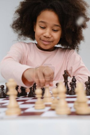 Téléchargez les photos : Afro-Américaine jouant aux échecs. Joyeux enfant souriant derrière les échecs souriant en classe ou à l'école leçon. excité intelligent noir gosse avec jeu de société close-up - en image libre de droit