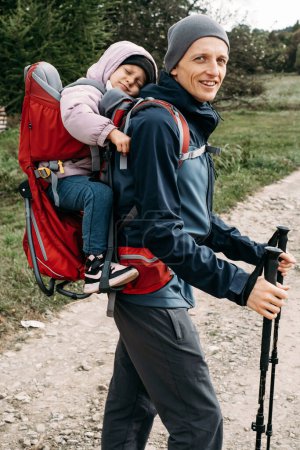 Foto de Hombre viajero con niño cansado en la mochila Padre lleva a su hija en una mochila Hombre llevó niño deslizante a las montañas. Viajar con niños. Caminando con el bebé en otoño, paseo nórdico - Imagen libre de derechos