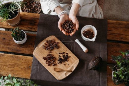 Foto de Manos de mujer sosteniendo granos de cacao orgánicos en mesa de madera, plumas de cacao, fabricación de chocolate artesanal en estilo boho rústico para la ceremonia. Degustación, fabricación de chocolate con vista superior de primer plano - Imagen libre de derechos