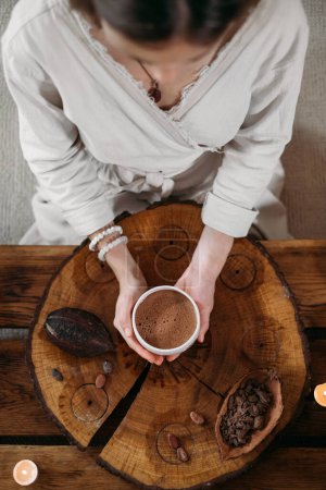 Foto de Cacao ceremonial caliente hecho a mano en taza blanca. Manos de mujer sosteniendo cacao artesanal, vista superior sobre mesa de madera. Bebida orgánica saludable de chocolate preparada a partir de frijoles, sin azúcar. Dando taza en la ceremonia, café acogedor - Imagen libre de derechos