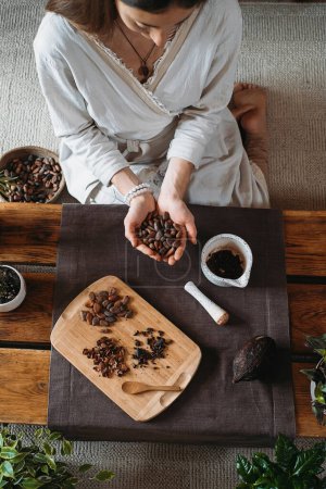 Photo for Woman hands holding organic cacao beans on wooden table, cocoa nibs, artisanal chocolate making in rustic boho style for ceremony. Degustation, Chocolate making with pounder close-up top view - Royalty Free Image