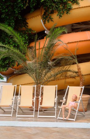 deck chairs in indoor water park for rest near palm trees. Child sitting near slide in aquapark, peace. Copy space.
