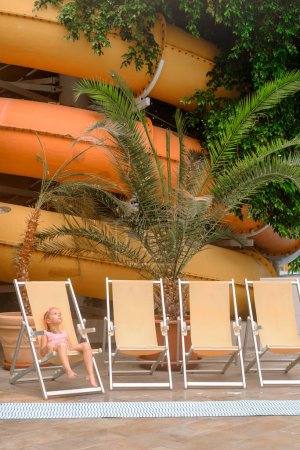 Liegestühle im überdachten Wasserpark zum Ausruhen in der Nähe von Palmen. Kind sitzt neben Rutsche im Aquapark, Ruhe. Kopierraum.