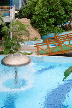 Water fountain in aquapark. Mushroom shape fountain in indoor adventure park full of green palm trees with bridge and swimming pools, hydro massage tubs indoor