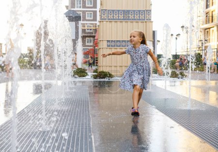 Téléchargez les photos : Météo estivale chaude. Enfant jouant dans une fontaine d'eau dans la ville. Fille heureuse jouant avec des arroseurs d'eau, courant et riant. Enfance active. Jeux en plein air. Rue de la ville moderne - en image libre de droit