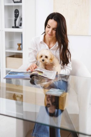 Foto de Mujer sentada junto a la mesa con maltipoo pequeño perro descansando en sus regazos, leyendo revista en la oficina - Imagen libre de derechos