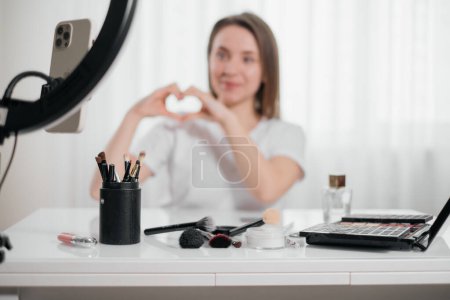 Foto de Blogger mostrando cosméticos y forma de corazón Mujer haciendo maquillaje en casa, aplicar cosméticos decorativos. Hermosa joven hembra usando cepillo cerca del espejo y el teléfono inteligente. - Imagen libre de derechos