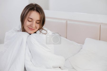 Téléchargez les photos : Jolie jeune femme caucasienne souriante se réveillant le matin et s'étirant dans son lit et regardant contenu. Couvert de lin blanc. - en image libre de droit