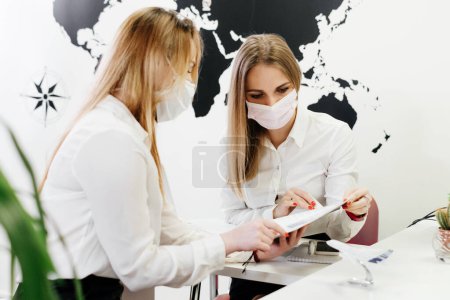 Photo for Travel agent having a client in light modern office of tour agency, choose destination. Assistant speaking in face mask in the travel agency to clients. protection during covid pandemic - Royalty Free Image