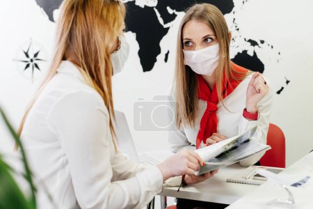 Photo for Travel agent having a client in light modern office of tour agency, choose destination. Assistant speaking in face mask in the travel agency to clients. protection during covid pandemic - Royalty Free Image