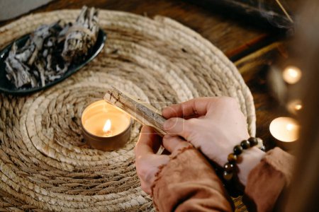 Photo for Woman hands burning Palo Santo, before ritual on the table with candles and green plants. Smoke of smudging treats pain and stress, clears negative energy and meditation wooden stick - Royalty Free Image