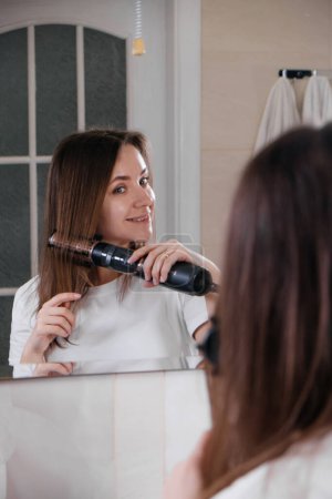 Téléchargez les photos : Femme séchant ses cheveux, coiffant dans la salle de bain, regardant le miroir, peignant les cheveux à la maison - en image libre de droit