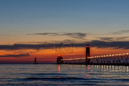 Téléchargez les photos : Un voilier s'approche du phare de Grand Haven, Michigan, au coucher du soleil sur le lac Michigan. Photo de haute qualité - en image libre de droit