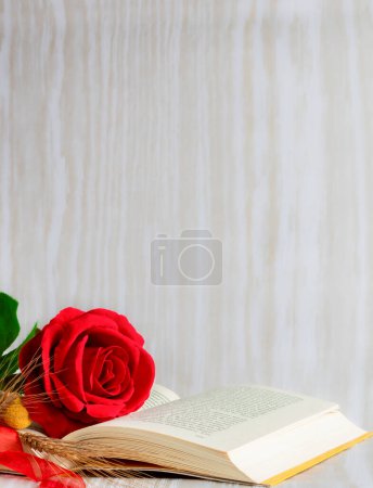 Foto de Rosa roja y libro abierto con cubiertas amarillas, espiga de trigo y lazo rojo sobre fondo textura clara, fondo vertical, beige para escribir - Imagen libre de derechos