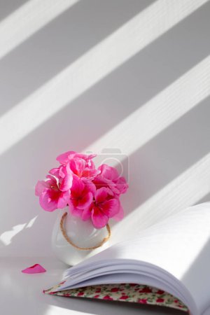 Téléchargez les photos : Petit vase blanc avec géraniums roses et un livre ouvert avec des feuilles blanches, avec un fond blanc et grisâtre avec les lignes des stores. Vertical. - en image libre de droit