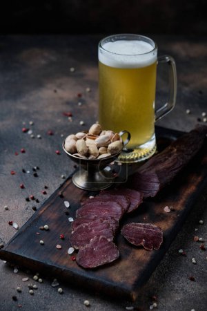 Photo for Homemade jerky meat on a kitchen cutting board and glass of beer on dark wooden  background - Royalty Free Image