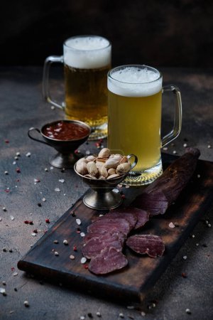 Photo for Homemade jerky meat on a kitchen cutting board and two glasses of beer on dark wooden  background - Royalty Free Image