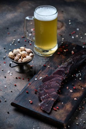 Photo for Homemade jerky meat on a kitchen cutting board with glass of beer on dark wooden  background - Royalty Free Image