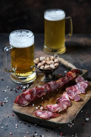 Photo for Cutting Spanish sausage fuet on a kitchen cutting board and two glasses of beer on dark wooden  background - Royalty Free Image