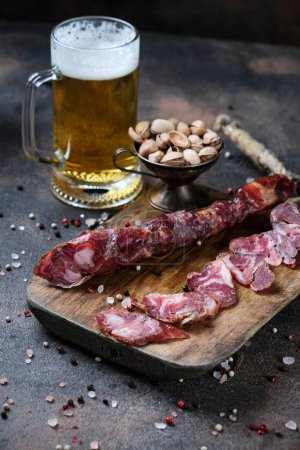 Photo for Cutting Spanish sausage fuet on a kitchen cutting board and glass of beer on dark wooden  background - Royalty Free Image