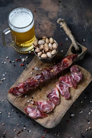 Photo for Cutting Spanish sausage fuet on a kitchen cutting board and glass of beer on dark wooden  background - Royalty Free Image