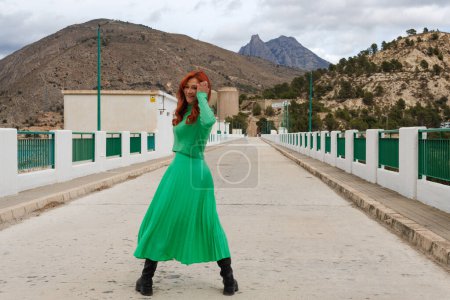 Reife Frau mit grünem Kleid und roten Haaren an der Wand des Amadorio-Stausees und dem Puig Campana im Hintergrund. Orcheta, Spanien