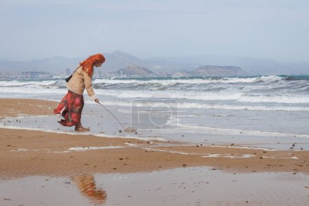 Red-haired woman poking a jellyfish with a stick to check if it still lives on El Altet beach, Spain