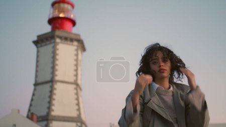 Beautiful girl resting lighthouse view. Casual young woman looking camera adjust curly hair. Worried pensive female contemplating walking seaside. Serious traveler turning enjoying evening trip alone