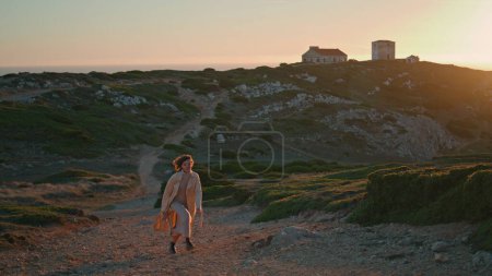 Cheerful woman walking sunset pathway on ocean shore cliff. Happy tourist admire landscape strolling rocky road mountain at seaside. Beautiful curly traveler enjoy evening. Weekend leisure concept.