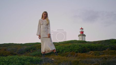 Photo for Blonde model posing nature green hill. Cute woman watching camera taking different positions at lighthouse background. Calm girl in naked back dress standing sunset environment. Tender lady alone - Royalty Free Image