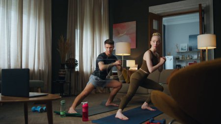 Photo for Fit man workouting with slim woman in room watching video lesson. Sporty family couple training body on mat at home. Young guy exercising with blond girl stretching legs. Fitness practice together - Royalty Free Image