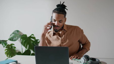 Foto de Positive freelancer talking phone in light office closeup. African american man working laptop answering call at home workplace. Hipster guy having conversation remotely. Involved specialist chatting - Imagen libre de derechos