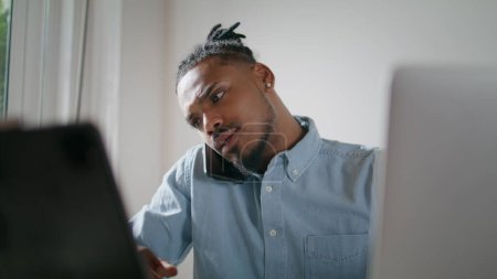 Foto de Busy freelancer talking phone in light office closeup. African american man working laptop answering call at home workplace. Hipster guy having deadline remotely. Involved specialist solving problems - Imagen libre de derechos
