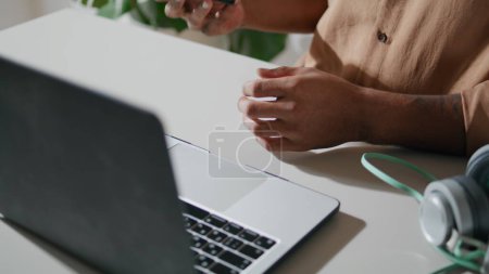 Foto de Focused businessman calling cellphone in office closeup. Young man answering phone call at home moving shot. Unshaven african american guy using smartphone. Modern freelancer sitting at laptop desk - Imagen libre de derechos