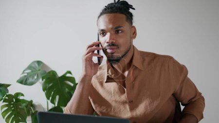 Foto de Busy man chatting mobile in office closeup. Afro american freelancer working laptop ending call at home workplace. Black hair guy picking down phone working remotely. Focused hipster typing keyboard - Imagen libre de derechos