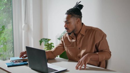 Foto de Focused man taking glasses on in office closeup. Dreadlocks guy working laptop at home remotely. Serious freelancer looking computer screen preparing taking notes. Afro american student sitting alone - Imagen libre de derechos