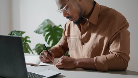 Foto de Focused freelancer writing notes in office closeup. Dreadlocks guy working at laptop table. Serious man studying attentively at computer desk. Afro american student sitting alone at home remotely - Imagen libre de derechos