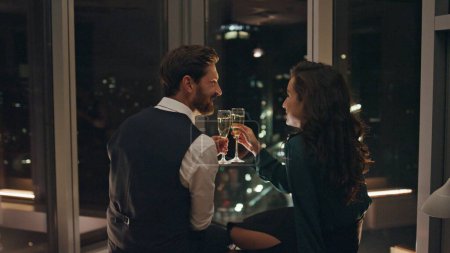 Photo for Attractive bearded man flirting with smiling woman drinking champagne late evening indoors. Two lovers relaxing together enjoying romance. Happy couple have pleasant talk looking on night city view. - Royalty Free Image