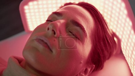 Mujer atractiva relajándose bajo la luz led en el procedimiento fotodinámico de cerca. Cliente clínico acostado en fototerapia no invasiva rejuvenecimiento facial eficaz e indoloro. Concepto de medicina estética.