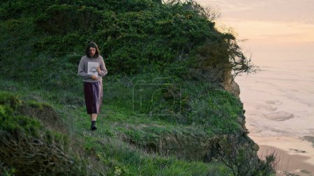 Lonely thoughtful woman walking coast hill at gloomy evening weather holding album with drawings. Pensive relaxed girl stepping on green grass seashore alone. Travel loneliness calmness concept.