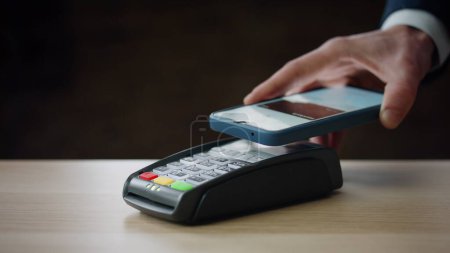 Foto de Cashless payment using smartphone on modern bank terminal indoors close up. Unknown person making purchase with contactless nfc technology. Man hand holding telephone for pay pass transaction. - Imagen libre de derechos