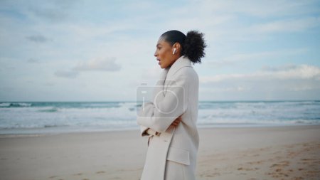 Téléchargez les photos : Ocean woman parler téléphone écouteurs sans fil sur le pouce. Afro-américain profiter de la promenade sur la plage bavarder avec un ami. Femme d'affaires paisible cheveux noirs travaillent à distance parlant cellule. Week-end concept de loisirs. - en image libre de droit
