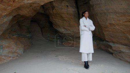 Unhappy girl thinking troubles at seashore cave. Lonely woman with crossed hands contemplating looking camera at beach cliff. Serious worried african american feel despair. Melancholy solitude concept